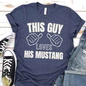 May include: A navy blue t-shirt with the text "This Guy Loves His Mustang" in white lettering. The shirt features two white thumbs up symbols on either side of the text.