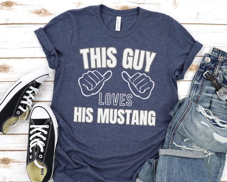 May include: A navy blue t-shirt with the text "This Guy Loves His Mustang" in white lettering. The shirt features two white thumbs up symbols on either side of the text.