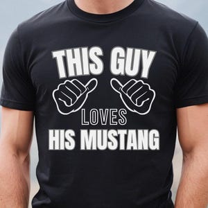 May include: Black t-shirt with white text that reads "This Guy Loves His Mustang". The text is surrounded by two white thumbs up symbols.
