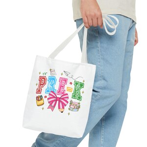 May include: A white canvas tote bag featuring a colorful graphic print that reads PRE-K in block letters. The design includes illustrations of a pink striped bow, a pencil, a box of crayons, a stack of books, a paper airplane, and small stars.