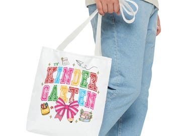 May include: A white tote bag featuring the colorful text KINDERGARTEN in a varsity font. The design includes a pink and white striped bow, a yellow crayon box, a stack of books, a paper airplane, and a small note with hearts on the bag.