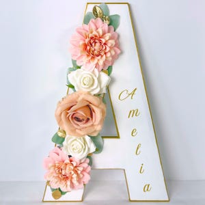 May include: A white letter A with gold trim, decorated with artificial peach and white flowers and green leaves. The name Amelia is printed in gold cursive script vertically along the right side of the letter. The design features layered blooms.