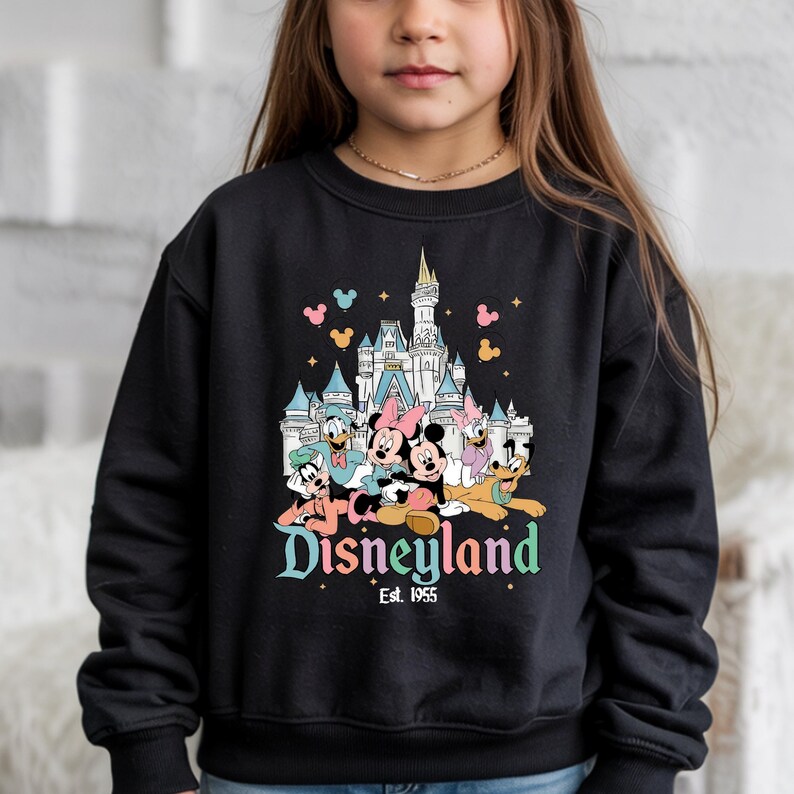 Puede incluir: Sudadera negra con un gr&aacute;fico colorido de personajes de Disney frente a un castillo. La palabra "Disneyland" est&aacute; escrita en una fuente caprichosa, con "Est. 1955" debajo. El dise&ntilde;o incluye Mickey y Minnie Mouse, el Pato Donald, Daisy Duck y Goofy.