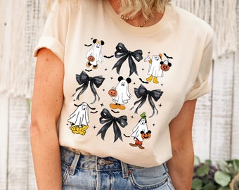 Puede incluir: Una camiseta de color crema con un diseño de temática de Halloween que presenta fantasmas de dibujos animados vestidos como Mickey Mouse, Minnie Mouse y Donald Duck. Los fantasmas sostienen calabazas y hay lazos negros dispersos por todo el diseño.