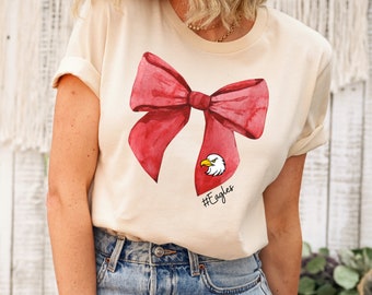 May include: A cream-colored t-shirt with a large red watercolor bow and an eagle graphic. The text "#Eagles" is printed below the eagle.