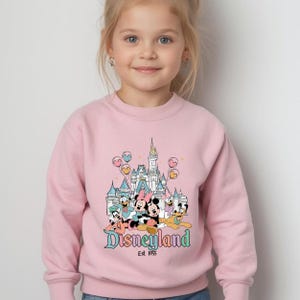 Puede incluir: Sudadera rosa claro con un gr&aacute;fico de Disneyland. El gr&aacute;fico incluye un castillo, Mickey y Minnie Mouse, el Pato Donald y Goofy. Tambi&eacute;n se ve el texto "Disneyland Est. 1955".