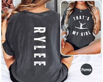 以下が含まれることがあります： 黒のTシャツで、背面に白い文字で「RYLEE」と印刷されています。Tシャツの前面には、体操選手の白いグラフィックと、その下に「THAT'S MY GIRL」という文字が印刷されています。