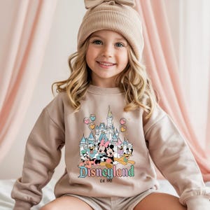 Puede incluir: Una sudadera beige con un gr&aacute;fico de Disneyland que presenta a Mickey, Minnie, Donald y Pluto, adem&aacute;s de un castillo y el texto "Disneyland Est. 1955". La sudadera la lleva una ni&ntilde;a peque&ntilde;a que lleva un gorro beige a juego.