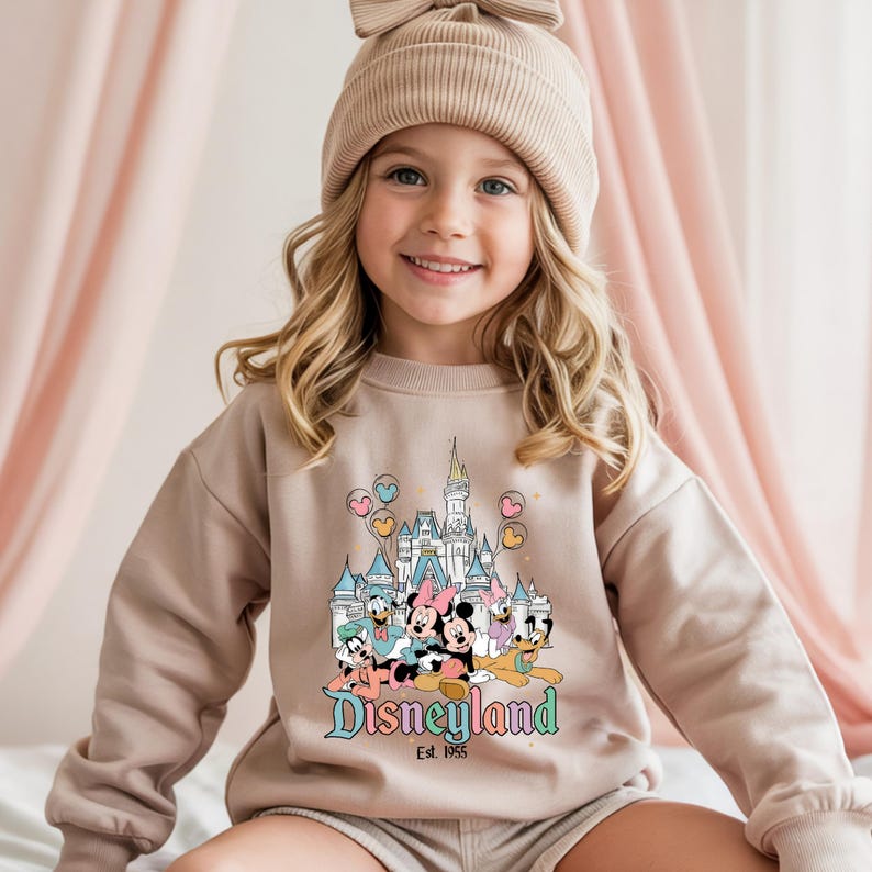 Puede incluir: Una sudadera beige con un gr&aacute;fico de Disneyland que presenta a Mickey, Minnie, Donald y Pluto, adem&aacute;s de un castillo y el texto "Disneyland Est. 1955". La sudadera la lleva una ni&ntilde;a peque&ntilde;a que lleva un gorro beige a juego.
