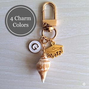 May include: Gold-colored keychain with a lobster clasp, featuring a white and gold initial charm, a graduation cap charm with "2026", and a seashell. The text "4 Charm Colors" is in a gray circle.