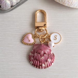 May include: A gold-colored keychain with a lobster clasp, featuring a pink heart charm with "love" text, a white circle with the letter "S", a pearl, and a pink and white seashell charm.