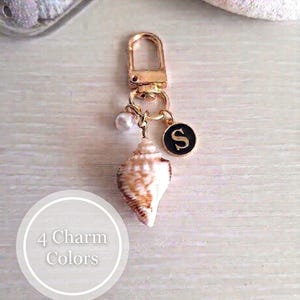 May include: Gold-colored keychain with a seashell charm, pearl, and a black circular charm with the letter "S". The seashell is brown and white. The image also includes text that says "4 Charm Colors".