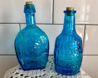 Peut inclure: Deux bouteilles en verre turquoise avec bouchons en liège. Les bouteilles présentent des motifs en relief et sont présentées sur un napperon en crochet blanc. L'une est arrondie, l'autre cylindrique.