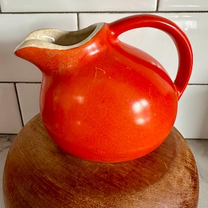 Könnte beinhalten: Ein leuchtend orangefarbener Keramikkrug mit rundem Körper und gebogenem Henkel. Der Krug hat einen Ausguss und einen cremefarbenen Rand. Er steht auf einer Holzschale. Der Krug wird wahrscheinlich zum Servieren von Getränken verwendet.