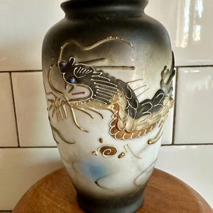 May include: A white ceramic vase with a black and gold dragon design. The dragon has blue eyes and is surrounded by gold and black accents.