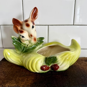 May include: A vintage ceramic planter featuring two deer figures nestled among green leaves. The planter has a light yellow base with a central opening and is accented with red berries. The deer have brown and white spotted fur.