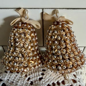May include: Two gold-colored bell ornaments, each adorned with sequins and faux pearls. The bells have cream-colored ribbon bows at the top and hang from a pearl-tipped loop. They rest on a white crocheted surface.