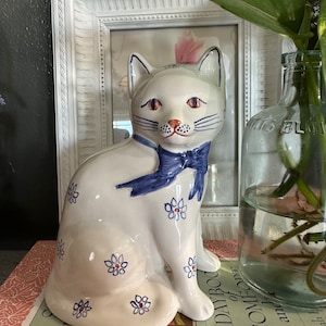 Könnte beinhalten: Weiße Keramik-Katzenfigur mit blauen und roten Blumenmustern und einer blauen Schleife. Die Katze sitzt auf einem Buch mit dem Text "A Daybook of Comedies and Jokes".