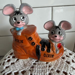 May include: A ceramic figurine featuring two cartoon mice in a brown boot. The mice have gray fur, pink ears, and are wearing a blue bow tie and a red bow tie. The boot has the word "Dixie" on it.