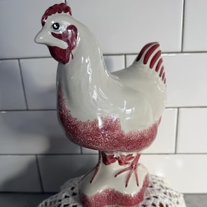 May include: A ceramic chicken ornament with a white body and red detailing. The chicken features a detailed head, comb, and wattle. The base is white with red accents. This figurine is likely a decorative item for a kitchen or farmhouse setting.