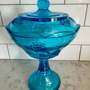 Könnte beinhalten: Eine türkisfarbene Bonbonniere aus Glas mit Deckel. Die Schale hat ein Weinrebenmuster und einen dekorativen Knauf auf dem Deckel. Die Schale besteht aus transparentem Glas.