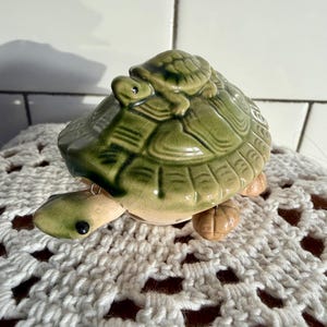 以下が含まれることがあります： 小さなカメが背中に乗った、セラミック製のカメの置物。置物は主に緑色で、足と頭にタン色のアクセントがあります。カメは白いかぎ針編みの表面に座っています。