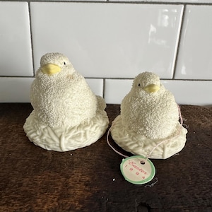 May include: Two white ceramic birds with yellow beaks sitting in nests. The birds are covered in a white, fluffy material. A tag is attached to the smaller bird that reads "Easter 1999".