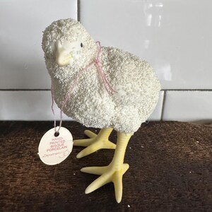May include: White porcelain chick figurine with yellow legs and feet. The chick is covered in white glitter and has a pink tag that reads "Hand Painted Bisque Porcelain *Department 56*"