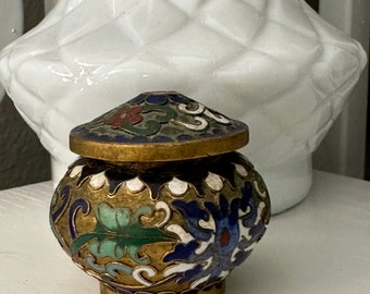 May include: A small, round, gold-colored trinket box with a lid. The box is decorated with blue, green, white, and red enamel floral designs.