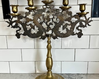 Puede incluir: Un candelabro de latón con seis candeleros. El candelabro presenta un diseño ornamentado con dos dragones y motivos florales. La base es redonda con detalles florales grabados. La altura total es de aproximadamente 38 cm.