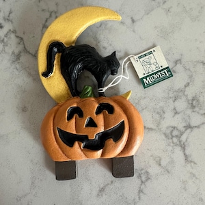 May include: A Halloween-themed decoration featuring a black cat perched on a yellow crescent moon above an orange jack-o'-lantern. The jack-o'-lantern has a carved face. The decoration has a tag that says "Midwest".