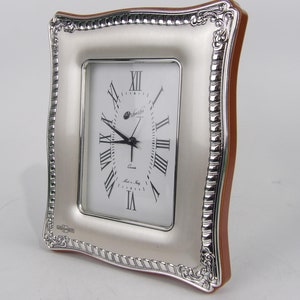 Może przedstawiać: Prostokątny zegar w kolorze srebrnym z białą tarczą i czarnymi rzymskimi cyframi. Zegar ma ozdobną srebrną ramę z brązowym drewnianym tyłem. Na tarczy widnieje napis "Quartz" i "Made in Italy".