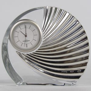 May include: A decorative desk clock with a clear, curved glass frame. The clock face is white with black numerals and hands. The back of the clock is a silver, fan-shaped design. The clock is approximately 13 cm tall.