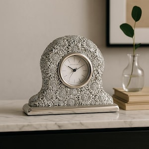 May include: Silver-toned decorative clock with a floral design. The clock face is white with black hands and numerals. The clock is on a rectangular base and has the word "PRIARCARY" printed on the clock face.