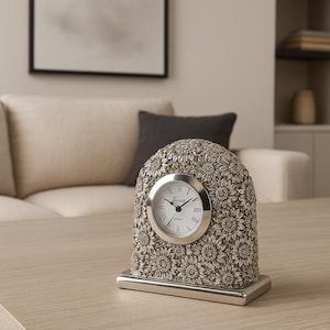May include: A decorative silver-colored desk clock with a floral design. The clock face is white with black hands and Roman numerals. The clock is on a rectangular base and sits on a light-colored wooden surface.