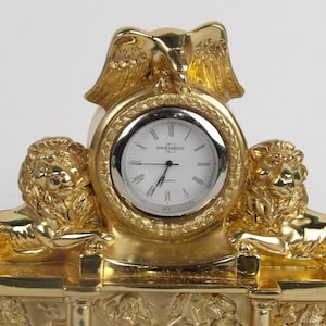 May include: A gold-coloured decorative clock with a white clock face and Roman numerals. The clock is flanked by two lion figures and topped with an eagle. The clock face reads "Lina Argenis".