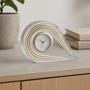 May include: A clear, teardrop-shaped desk clock with gold-coloured accents and a white clock face. The clock features the text "ILNO ARCENTE" and is set on a light-coloured wooden surface.