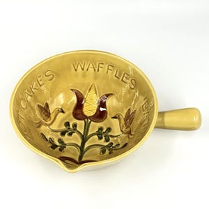 May include: A yellow ceramic batter bowl with a handle and a pouring spout. The interior features a brown and green floral design with two birds and the embossed text HOT CAKES, WAFFLES, and BATTER arranged around the inner rim of the bowl.