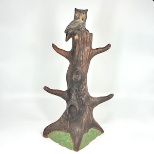 May include: A brown tree-shaped mug rack featuring a textured bark finish and four protruding branches for hanging items. A small, detailed owl with yellow eyes and grey and brown feathers is perched at the very top of the central trunk.