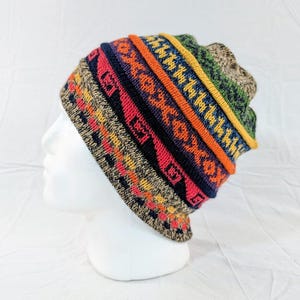 Könnte beinhalten: Eine bunte Strickmütze mit einem gemusterten Design. Die Mütze hat Bänder in Orange, Rot, Blau und Grün mit geometrischen Mustern. Die Mütze wird auf einem weißen Schaufensterkopf präsentiert.