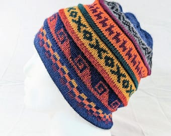 Può includere: Un berretto lavorato a maglia con un motivo geometrico colorato. Il cappello presenta una base blu con fasce arancioni, rosse, verdi e gialle. Il design include motivi ripetuti di diamanti e le lettere "x" e "o". Il cappello è esposto su una testa di manichino bianca.