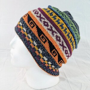Könnte beinhalten: Ein farbenfroher Strickbeanie mit geometrischem Muster. Der Hut hat Bänder in Orange, Lila, Gelb und Blau mit weißen und schwarzen Akzenten. Der Beanie wird auf einem weißen Schaufensterkopf präsentiert.