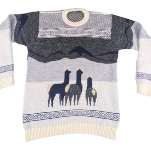 May include: A wool sweater with a mountain and llama design. The sweater features a cream-colored ribbed collar, cuffs, and hem. The body of the sweater has a gray and white landscape with three dark blue llamas. The sleeves are white with blue geometric patterns.