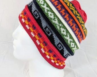 May include: A colorful knit beanie with a geometric pattern. The hat features bands of red, black, gray, green, white, orange, and purple. The beanie is displayed on a white mannequin head.