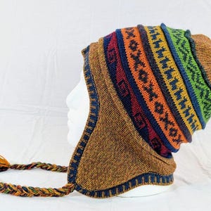 May include: A knitted hat with ear flaps and a braided tassel. The hat has a brown base and a geometric pattern in red, orange, blue, and green. The ear flaps are trimmed with dark blue.