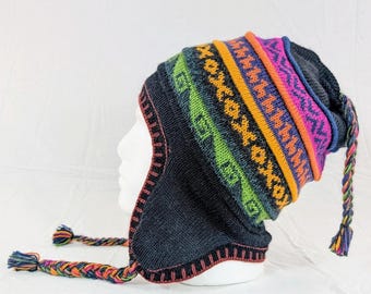 May include: A dark gray knit hat with ear flaps and a colorful geometric pattern. The hat features bands of orange, pink, blue, and green, with braided tassels in matching colors. The hat is displayed on a white mannequin head.