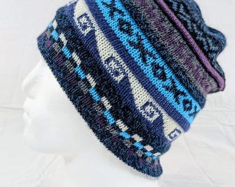 Può includere: Un berretto lavorato a maglia con un motivo geometrico colorato. Il cappello presenta sfumature di blu, bianco e viola. Il bordo è blu scuro e il bordo superiore è rifinito con una fascia viola. Il cappello è esposto su una testa di manichino bianca.