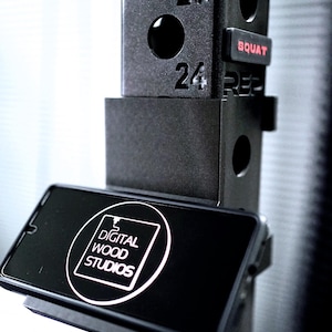May include: Black weightlifting rack with the numbers 24 and 25 visible. A phone is mounted on the rack, displaying the text "DIGITAL WOOD STUDIOS" in a white circle. The word "SQUAT" is in red.