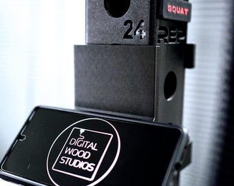 May include: Black weightlifting rack with the numbers 24 and 25 visible. A phone is mounted on the rack, displaying the text "DIGITAL WOOD STUDIOS" in a white circle. The word "SQUAT" is in red.