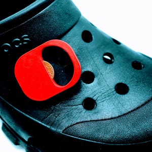 Puede incluir: Primer plano de un zapato Crocs negro con un accesorio rojo brillante adjunto. Se ve una moneda dentro de la abertura del accesorio. El zapato tiene varios agujeros redondos y se ve el nombre de la marca.